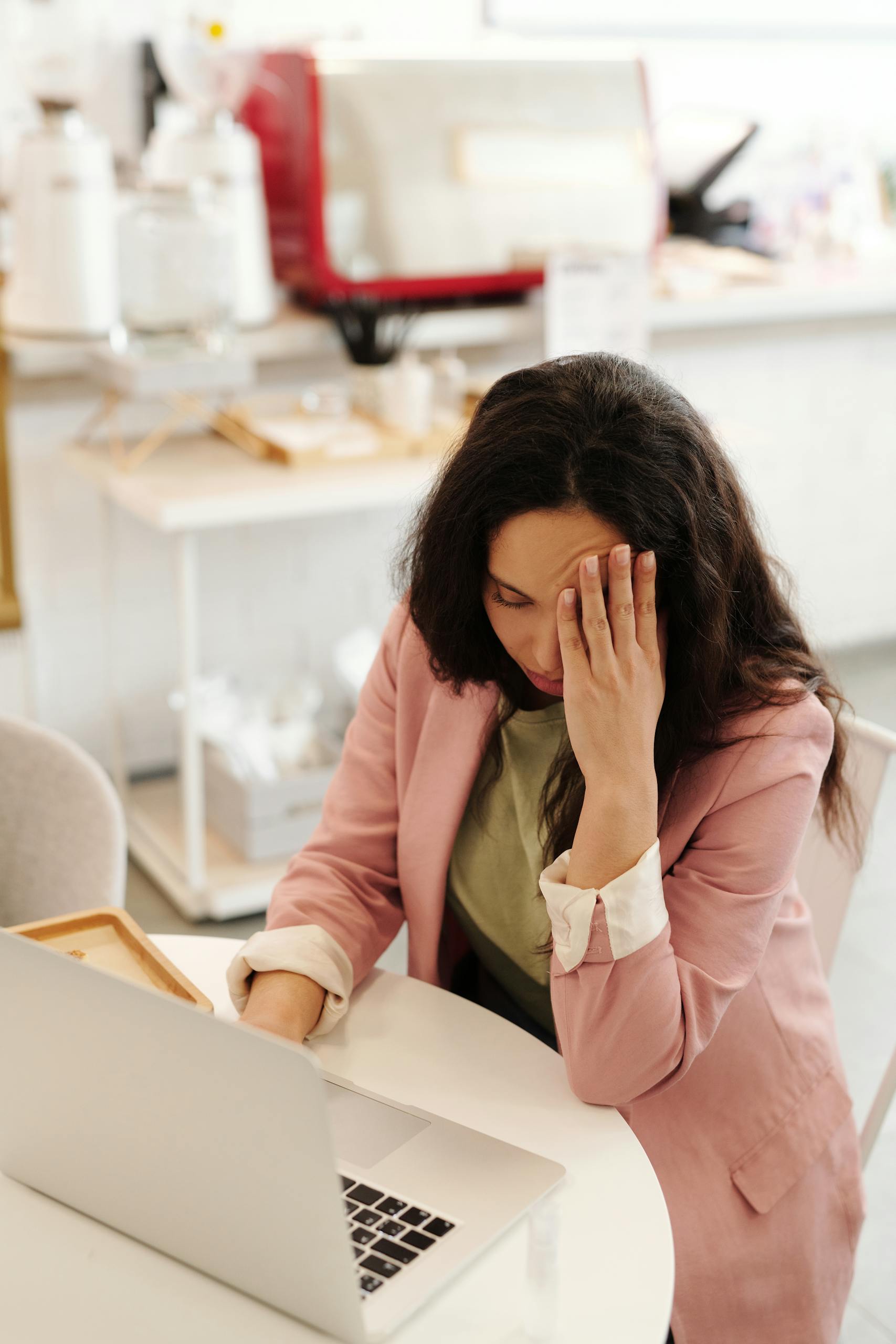 Woman holding head in hands while working on a laptop indoors, seemingly stressed or unwell.